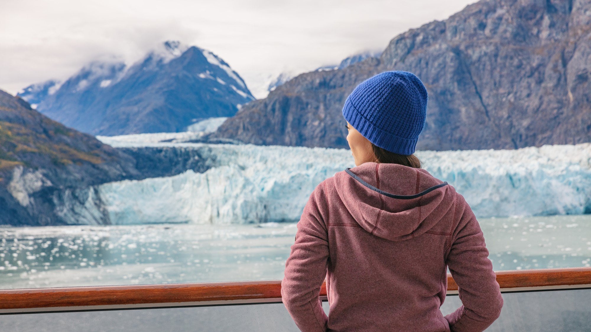 A woman standing on the deck of a cruise ship on an Alaskan cruise looking over the railing at a glacier